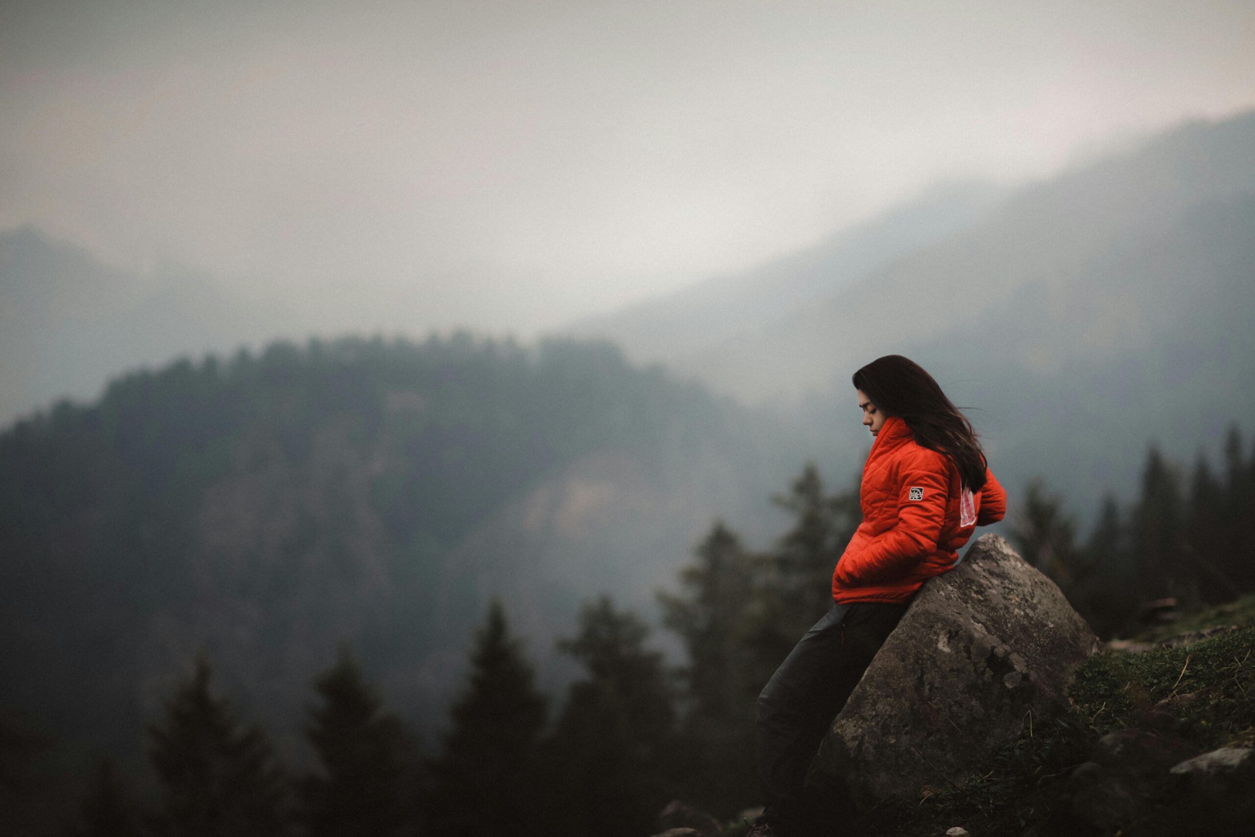 A woman in a red jacket leans against a boulder, enjoying the mountainous view in India.